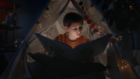 Excited boy with blond hair in makeshift hut opening book at home in evening. Teenager sitting on floor while reading book with flashlight. Concept of leisure and free time