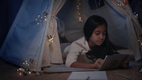 Cheerful girl spending time in decorative makeshift hut at home in evening. Pretty teen lying on floor while watching video on tablet. Concept of leisure and carelessness