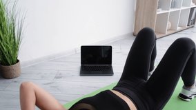 Fitness training. Athletic woman. Abs workout. Online sport. Healthy lifestyle. Sportive defocused lady doing abdominal crunches workout with laptop black screen in light room interior.