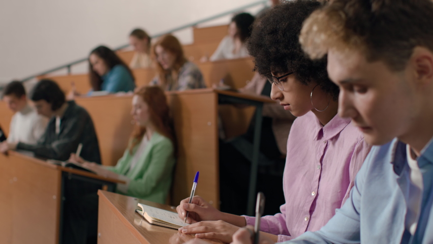 Group of students learns at lecture room. Young people study in university or pupil sitting at school lesson. Teens listen to teacher and write at auditorium. Modern education indoors of college hall | Shutterstock HD Video #1071999712