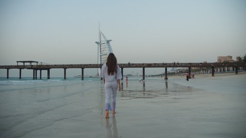 Back view of attractive young brunette woman with long hair walking on sand beach in Dubai towards Burj Al Arab hotel in Dubai, UAE in 2021