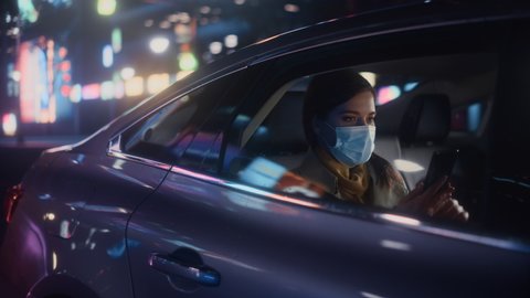 Female Wearing Face Mask is Commuting Home in Backseat of a Taxi at Night. Beautiful Passenger Using Smartphone and Looking Out of Window while in a Car in Urban City Street with Working Neon Signs.