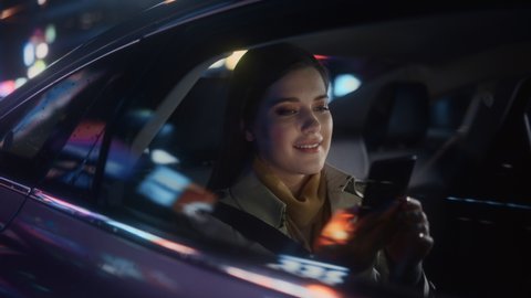 Happy Female is Commuting Home in a Backseat of a Taxi at Night. Beautiful Woman Passenger Making a Video Call on a Smartphone while in Transfer Car on City Street with Working Neon Signs.