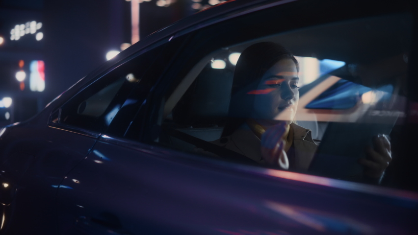 Stylish Female is Commuting Home in a Backseat of a Taxi at Night. Beautiful Woman Passenger Using Tablet Computer and Looking Out of Window while in Car in Urban City Street with Working Neon Signs. Royalty-Free Stock Footage #1072002667