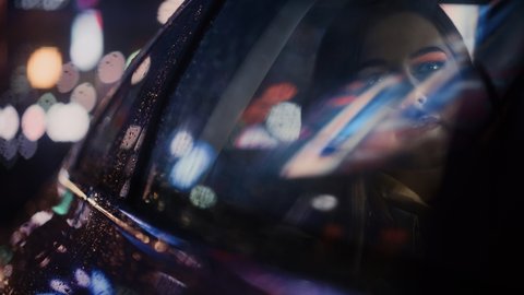 Stylish Female is Commuting Home in a Backseat of a Taxi at Night. Beautiful Woman Passenger Looking Out of Window while in a Car in Urban City Street with Working Neon Signs. Cinematic Footage.
