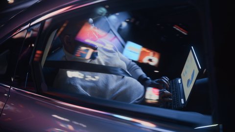 Stylish Black Man in Glasses is Commuting Home in a Backseat of a Taxi at Night. Male Using Laptop Computer and Looking Out of Window while in a Car in Urban City Street with Working Neon Signs.