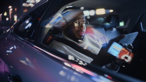 Stylish Black Man in Glasses is Commuting Home in a Backseat of a Taxi at Night. Male Using Tablet Computer and Looking Out of Window while in a Car in Urban City Street with Working Neon Signs. ஸ்டாக் வீடியோ