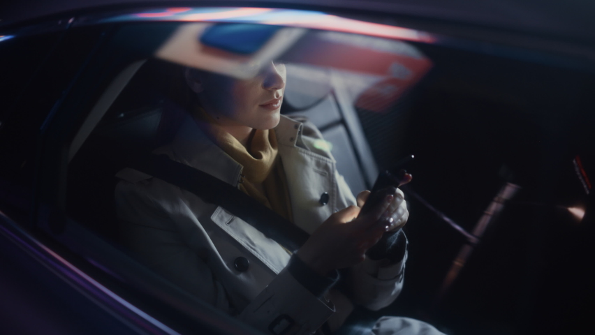 Stylish Female is Commuting Home in a Backseat of a Taxi at Night. Beautiful Woman Passenger Using Smartphone and Looking Out of Window while in a Car in Urban City Street with Working Neon Signs.