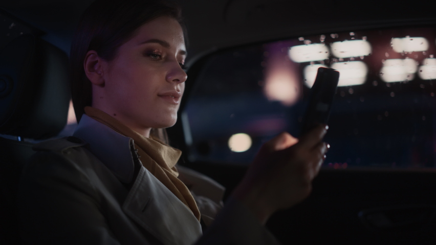 Stylish Female is Commuting Home in a Backseat of a Taxi at Night. Beautiful Woman Passenger Using Smartphone and Looking Out of Window while in a Car in Urban City Street with Working Neon Signs.