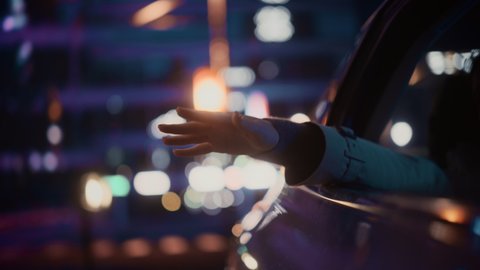 Female is Commuting Home in a Backseat of a Taxi at Night. Passenger Chilling and Holding Her Hand Outside of Window while in a Car in Urban City Street with Working Neon Signs. Cinematic Footage.