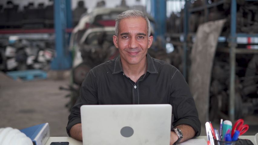 confident senior entrepreneur sitting on desk arms crossed  smiling with computer laptop looking at camera in warehouse of old used car engine . happy old businessman owner start up at garage store Royalty-Free Stock Footage #1072009804