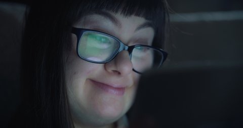 Cinematic shot of teen girl with down syndrome using tablet for entertainment while sitting on sofa at home at night. Concept of technology, connection, handicapped, disability, online, communication
