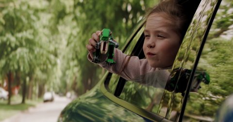 CU Portrait of cute little girl sticking head out of car window and playing with her toy dinosaur while riding through neighborhood. Shot with 2x anamorphic lens