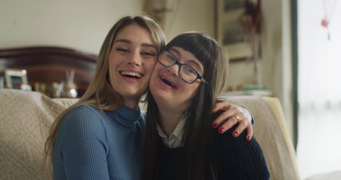 Authentic shot of happy young woman embracing her teen sister with down syndrome and smiling in camera at home. Concept of love, family, healthcare, persons with disabilities, friendship.