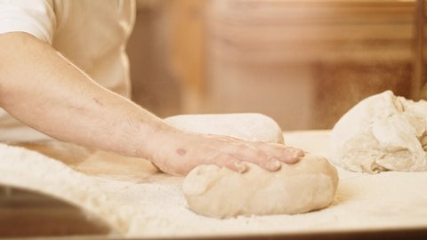 Close-up of hands of a male baker slamming dough on a table. Flour scatters in all directions, slomo, warm light, 4K, 