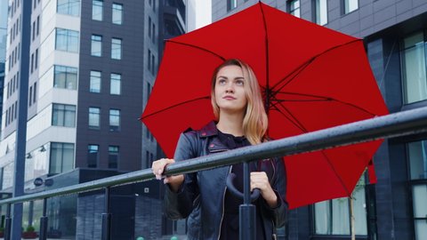 A portrait of a woman with red umbrella standing on the background of a modern building. A female model in black leather jacket is looking up thoughtfully