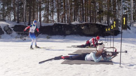 Professional biathletes lying in prone positions and shooting with rifles while their opponents skiing to shooting range