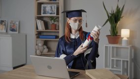 adult female graduate in academic clothes and mask on face rejoices in diploma while talking by video and on laptop during distance learning, takes off mask and looks at camera