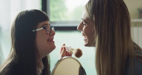 Cinematic shot of happy sister and teen girl with down syndrome are having fun to make up together in their room at home. Concept of love, family, persons with disabilities, beauty, friendship.