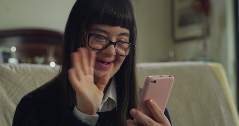 Cinematic shot of happy teen girl with down syndrome making video call with smartphone to friends or family at home. Concept of technology, persons with disabilities, media, friendship, new generation