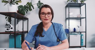 Happy beautiful 25-30 female doctor assistant in blue scrubs consulting remote patient online via webcam video chat app.