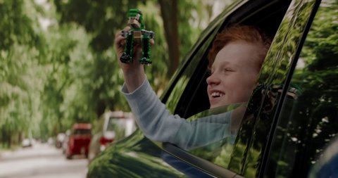 CU Portrait of cute little dreamer kid boy sticking head out of car window and playing with his toy dinosaur while riding through neighborhood. Shot with 2x anamorphic lens