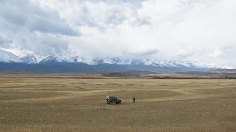Traveler with car observing mountains. Drone view of lonely anonymous adventurer standing near car amidst vast Altay steppe and camera moving towards mountains with snowy peaks under clouds