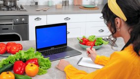 Blue screen mock up chroma key display laptop: Woman housewife in kitchen study watch online culinary course listen teacher tells chef writes culinary recipe in notebook, remote video call computer