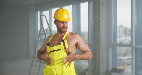 Sexy young shirtless builder in overall and hardhat dancing at construction site. Portrait of male stripper in construction worker uniform dancing in renovation house