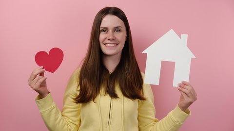 Portrait of friendly cute young woman 20s holding white paper house with red heart, happy looking at camera, wears hoodie, posing over pink background. Charity, real estate and family home concept