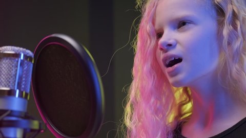 Portrait professional singer performs song singing into microphone in recording studio. Young blonde girl in recording studio sings song into professional microphone and records song.