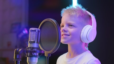 Portrait young blond boy singer wearing headphones, singing song in recording studio. Caucasian kid records new music track, sings song into microphone. Process of recording song in recording studio.