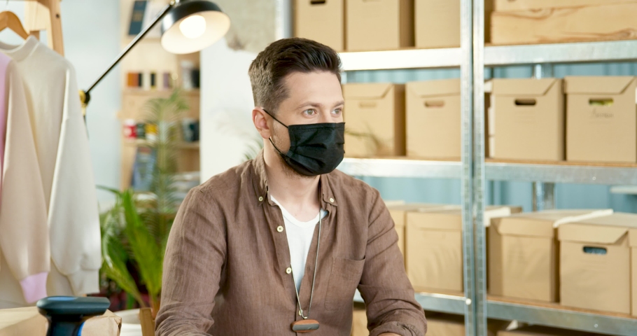 Close up portrait of Caucasian young joyful handsome male in black mask sitting at desk at workplace in clothing storehouse and looking at camera in good mood. Coronavirus quarantine