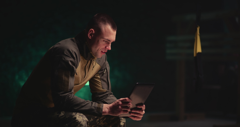 Pan left view of male soldier gesticulating and speaking with online friend while sitting and resting in dark gym of military base