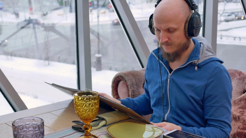 A bald young man with a well-styled beard and mustache, wearing black headphones, carefully leafs through the menu with pictures of dishes at the table. The table is prepared for the order.