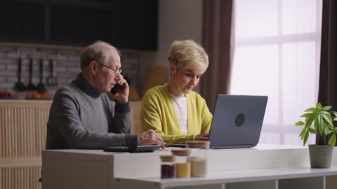 old man and woman are paying utility bills online, staying at home, husband is calling by phone and woman is using laptop with internet