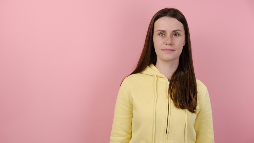 Excited young female showing aside with yellow paper arrow, dressed in hoodie, posing isolated over pink color background studio with copy space for advertisement. People emotion lifestyle concept
