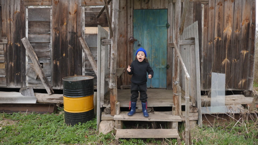 boy child in a black jumpsuit and a blue knitted hat stands on the porch