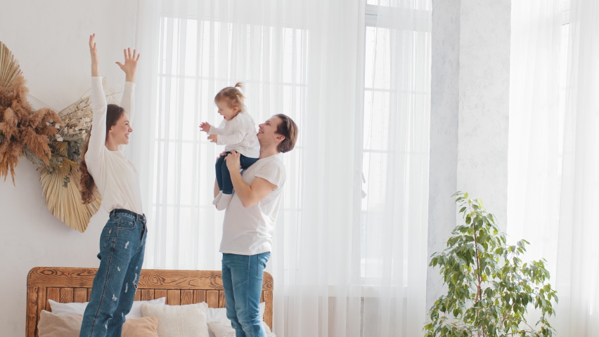 Parents young married couple caucasian family father holding little daughter baby girl lifts up in air mom raises hands child kid repeats, mum and dad smile enjoy happy weekend at home bedroom on bed Royalty-Free Stock Footage #1072070722