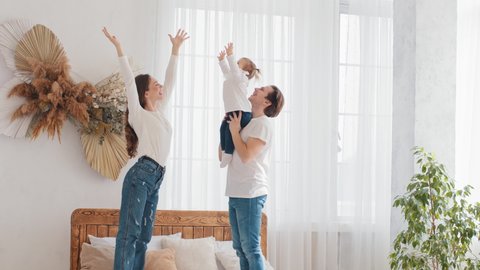 Parents young married couple caucasian family father holding little daughter baby girl lifts up in air mom raises hands child kid repeats, mum and dad smile enjoy happy weekend at home bedroom on bed
