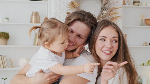 Portrait of caucasian family young parent holding little baby daughter toddler talking hugging smiling cuddling child girl showing direction with finger to side in window mom and dad looking away peep