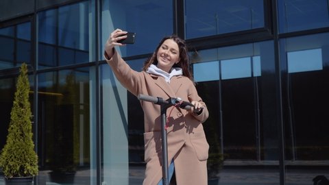 A young student girl in a sweater, jeans and a coat with an electric scooter stands near a stylish modern office center building and takes a selfie. Front view. Ecological city transport