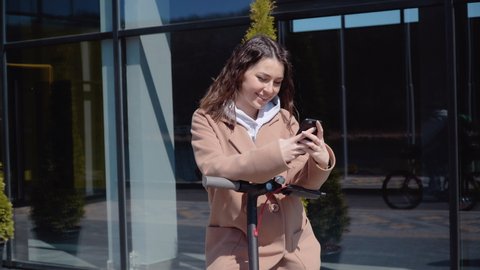 A young student girl in a sweater, jeans and a coat with an electric scooter stands near a stylish modern shopping center building and uses a smartphone. Front view