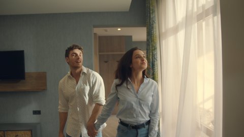 Front view of beautiful couple entering hotel room. Love couple walking in hotel room. Happy man and woman looking around in luxury suite. Smiling hipster couple falling back on bed indoors.