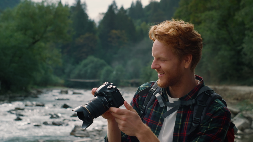 Happy guy with photo camera standing at river bank in mountains. Male photographer taking photos on professional camera. Young man looking pictures on digital camera outdoor Royalty-Free Stock Footage #1072080484