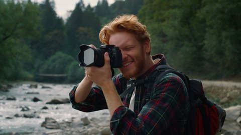 Smiling hiker shooting nature landscape in mountains. Young photographer using photo camera outdoor. Excited redhead man looking at photos on digital camera after shooting mountain river
