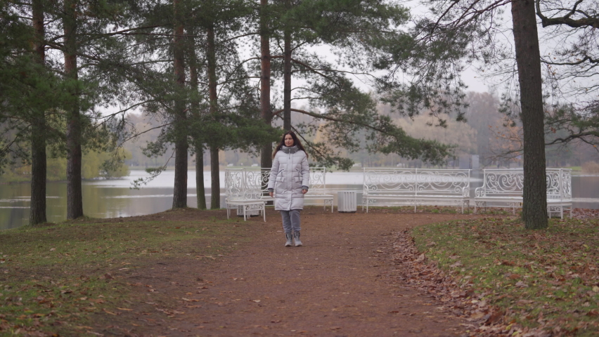 Young pregnant woman in warm coat walks on footpath in autumn park. Brunette woman with long hair in Catherine Park, Tsarskoye Selo, Saint Petersburg, Russia.