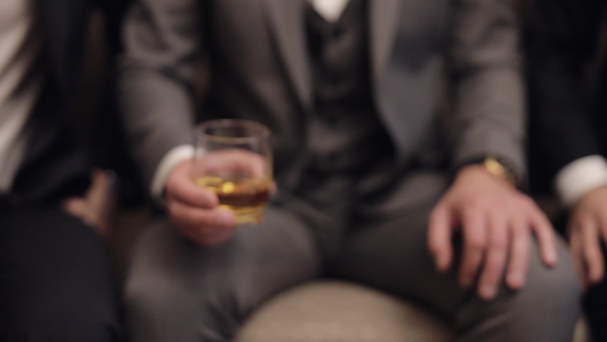 Company of grooms friends sitting, talking, holding and clinking glasses of alcoholic whiskey drinks in their hands. Male hands are holding glasses of cognac, brandy, close-up. Celebrate wedding day Royalty-Free Stock Footage #1072089511