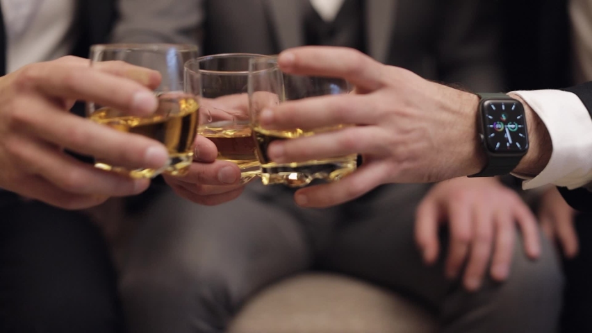Company of grooms friends sitting, talking, holding and clinking glasses of alcoholic whiskey drinks in their hands. Male hands are holding glasses of cognac, brandy, close-up. Celebrate wedding day | Shutterstock HD Video #1072089511