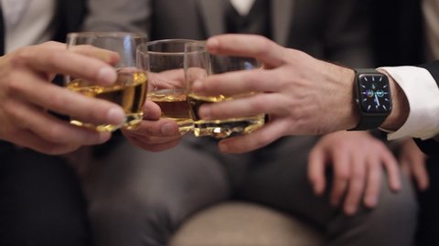 Company of grooms friends sitting, talking, holding and clinking glasses of alcoholic whiskey drinks in their hands. Male hands are holding glasses of cognac, brandy, close-up. Celebrate wedding day